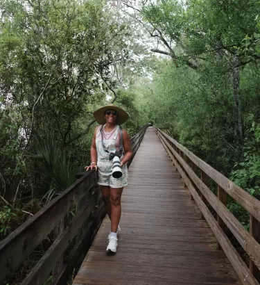 Smiling woman in a straw hat with a camera standing on a wooden boardwalk in the forest.
