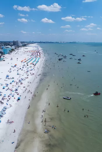 Aerial view of a white-sand beach lined with umbrellas and sunbathers, alongside shallow water dotted with small boats.