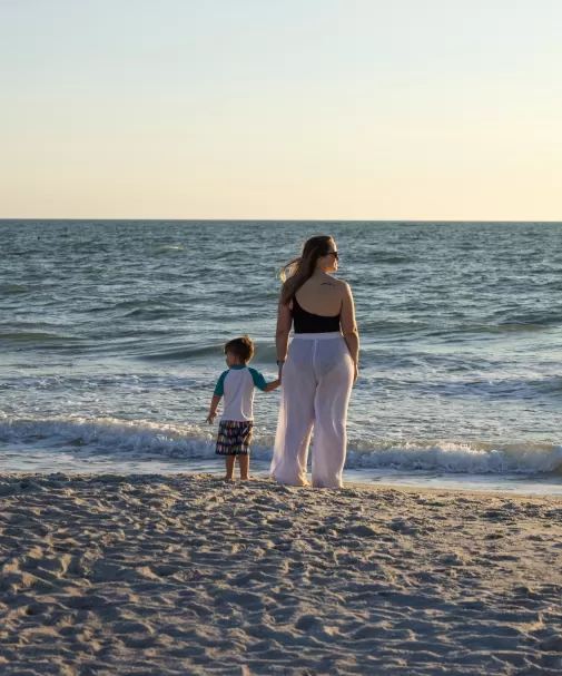 Woman and child holding hands at the water’s edge on a beach at sunset.